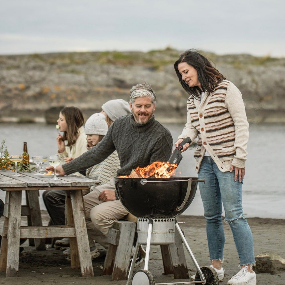 A group of people enjoying a barbecue by the beach, with one person using an Airlighter® to light the grill.