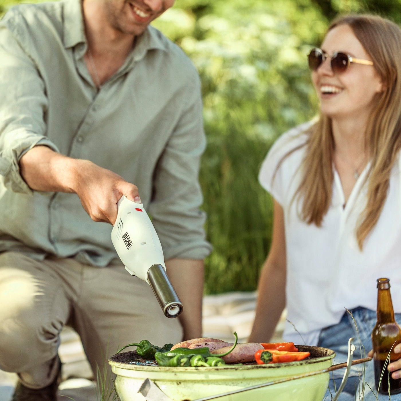 Man using a portable fire starter to ignite a grill while enjoying an outdoor BBQ with a smiling woman in a sunny, green setting.