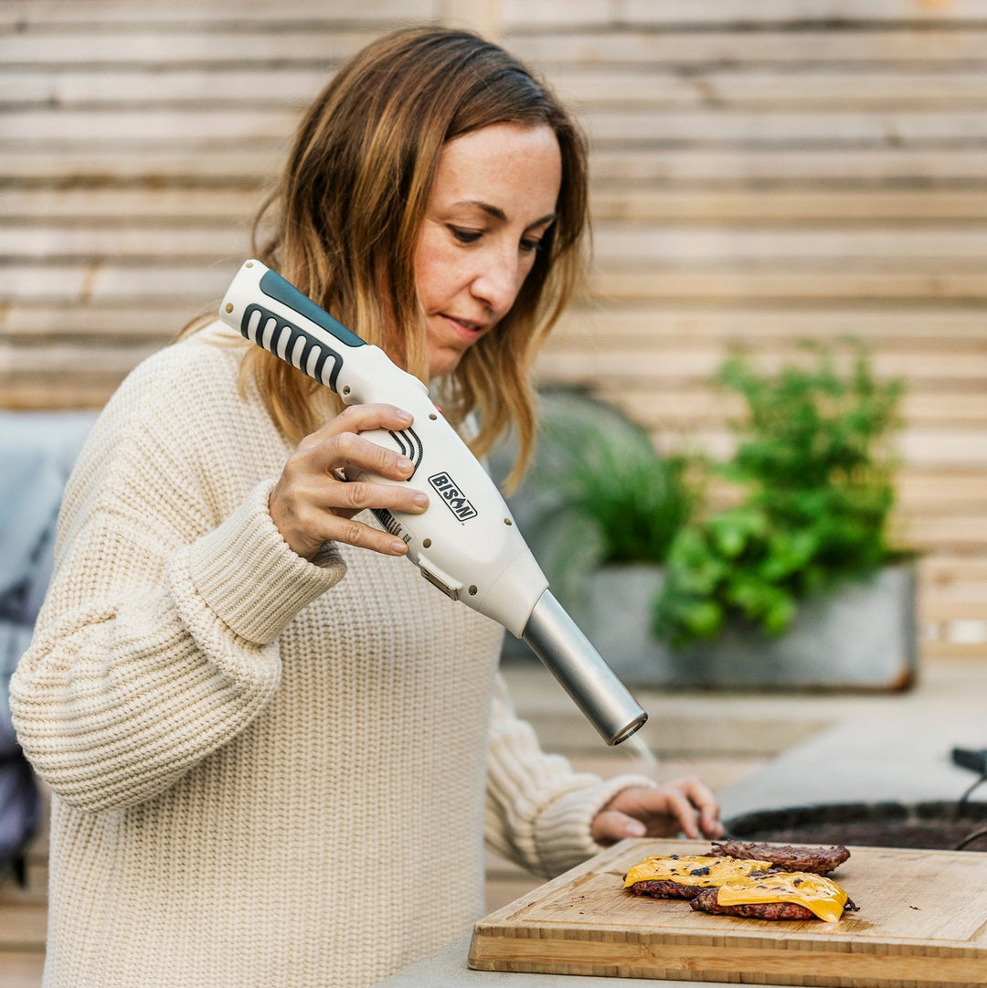 "Woman using a handheld culinary torch to sear cheeseburgers on a wooden cutting board outdoors."