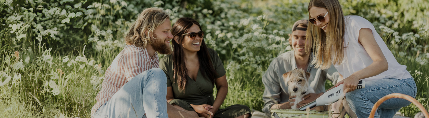 Group of friends enjoying a picnic in a lush green park, with one person using a portable grill and a small dog looking on.